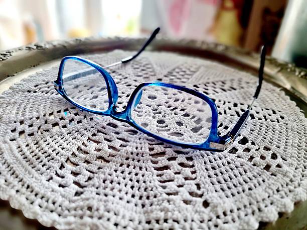 A pair of blue-rimmed glasses rests on a delicate white crocheted doily on a reflective surface