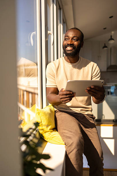 istock Man sitting by window holding tablet and smiling in sunlight 2212847666