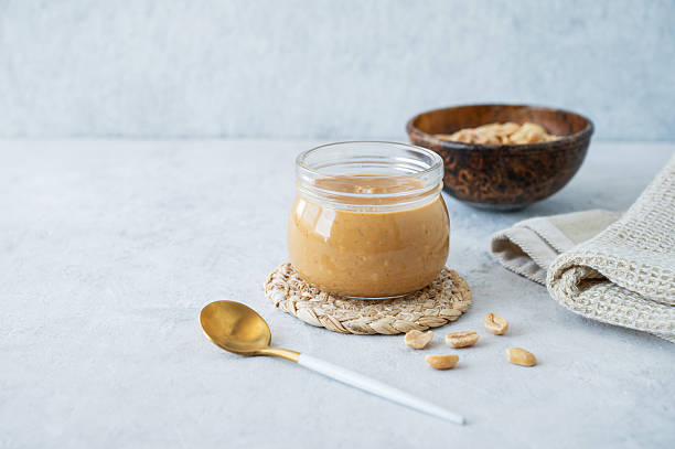 istock Peanut butter in glass jar on light background with salted nuts and napkin. 2212848458