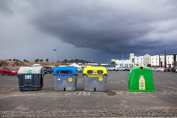 Arrecife, Lanzarote, Canary Islands, Spain - March 3rd 2025:  A row of waste sorting containers stands neatly arranged in a parking area, each designated for a specific recyclable material. From left to right: a gray bin for general waste, a blue container for paper and cardboard, a yellow bin for plastics, cans, and cartons, and a green bin for glass. Behind them, parked cars and buildings set the urban scene, emphasizing the role of structured recycling in public spaces. This image highlights sustainability, waste management, and environmental responsibility in an urban setting.