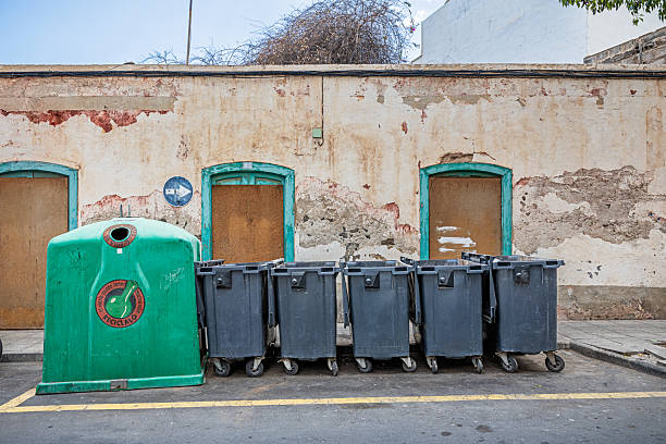 Arrecife, Lanzarote, Canary Islands, Spain - March 2nd 2025: A row of waste sorting containers stands neatly arranged in a parking area, each designated for a specific recyclable material. From left to right: a gray bin for general waste, a blue container for paper and cardboard, a yellow bin for plastics, cans, and cartons, and a green bin for glass. Behind them, parked cars and buildings set the urban scene, emphasizing the role of structured recycling in public spaces. This image highlights sustainability, waste management, and environmental responsibility in an urban setting.