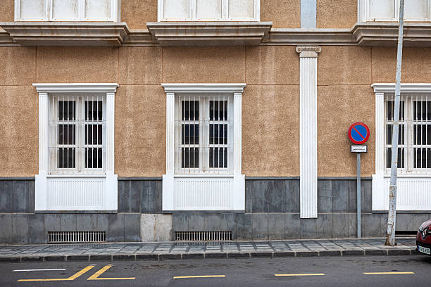 Arrecife, Lanzarote, Canary Islands, Spain - March 2nd 2025:  This image showcases a traditional Spanish facade with three symmetrically placed windows featuring white frames and protective bars. The textured beige wall with a gray stone base adds depth and authenticity, reflecting the architectural style commonly found in Lanzarote. A \