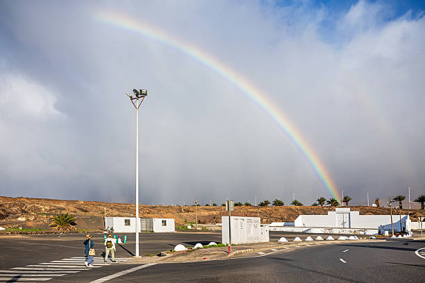 Arrecife, Lanzarote, Canary Islands, Spain - March 8rd 2025:  A vivid rainbow stretches across the sky, adding a splash of color to an otherwise muted urban scene. Below, a pedestrian crossing with bold white lines leads toward a traffic signal, where two individuals walk along the street. The towering streetlight and overhead power lines frame the composition, reinforcing the contrast between nature’s fleeting beauty and the structured city environment. The cloudy sky creates a moody atmosphere, making the rainbow’s presence even more dramatic.