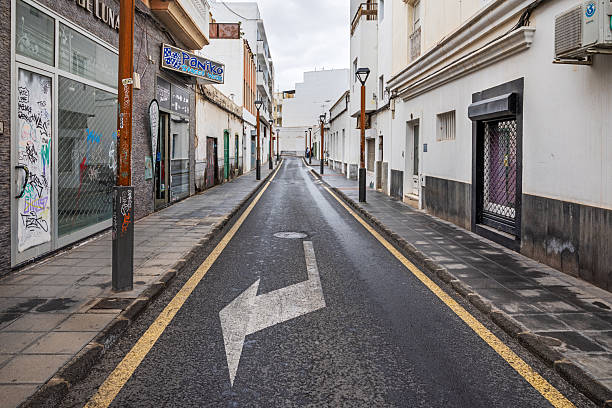 Arrecife, Lanzarote, Canary Islands, Spain - March 5th 2025:  This image captures a narrow one-way street framed by contrasting architectural styles. The worn facades of older buildings stand alongside modern structures, creating a visual dialogue between past and present. Yellow lines border the street, while a bold white \