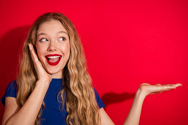 istock Young girl with charming expression presenting gesture against vibrant red background 2212854754