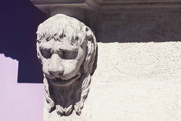 Stone carving of  lion head, with detailed features and fierce expression, adorns corner of a building's column. Sculpture casts a shadow on colored stone, with a purple wall visible in background