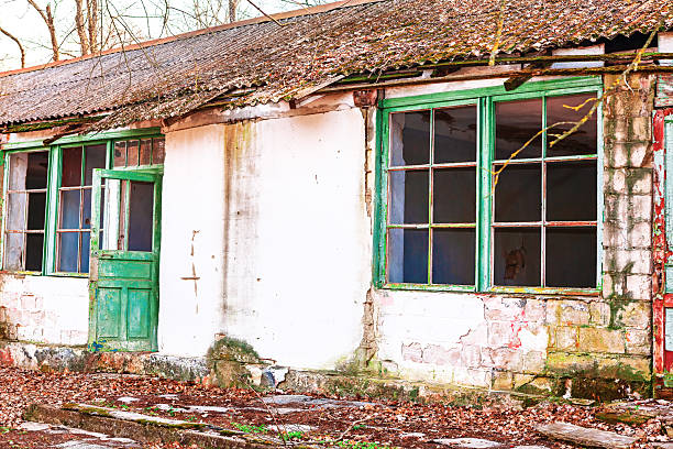Old abandoned building with green windows and door, showing significant signs of decay like peeling paint, moss growth, and broken glass. Weathered structure stands isolated, evoking forgotten stories