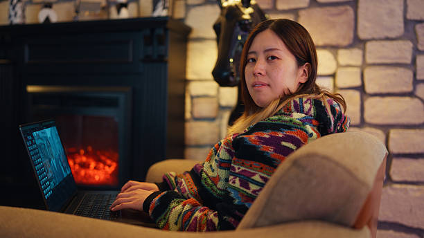 Portrait of woman sitting comfortably on a cozy sofa and working on her laptop by fireplace A portrait of a woman sitting comfortably on a cozy sofa and working on her laptop by a fireplace. female film editor stock pictures, royalty-free photos & images