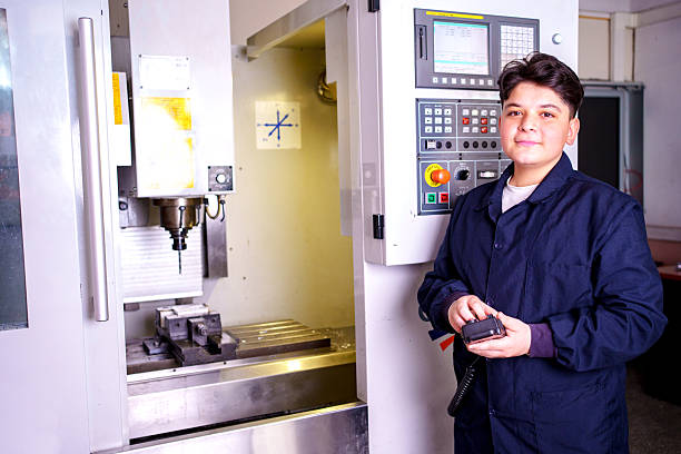 Confident young student in workwear posing next to a CNC (Computer Numerical Control) machine in a technical education center. Concept of digital manufacturing, modern machinery training, and vocational school.