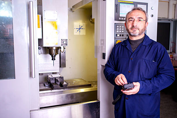 Confident mature adult worker in workwear posing next to a CNC (Computer Numerical Control) machine in a technical education center. Concept of digital manufacturing, modern machinery training, and vocational school.