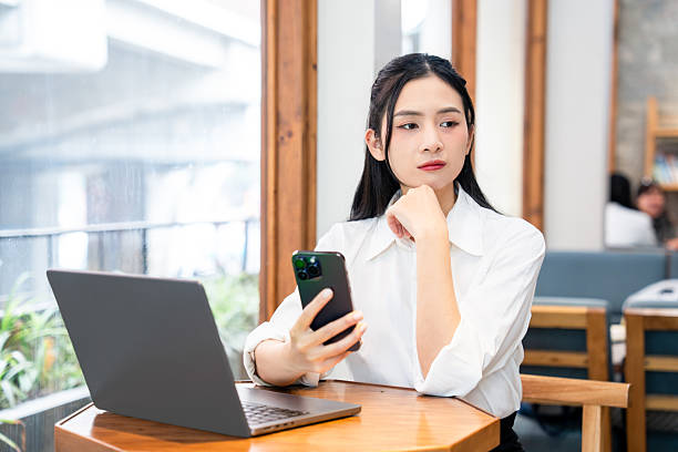 istock Young woman contemplates her phone while working on a laptop in a bright modern cafe setting 2212881587
