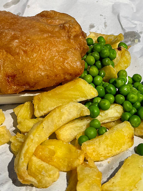 crispy fried fish fillet with mashed potatoes and green peas on white plate with fork, mustard and lemon slices on wooden table, vertical view from above