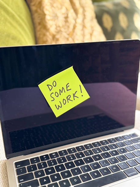 istock Image of yellow sticky note on screen of open laptop on sofa, 'Do Some Work!' written in black felt-tip pen, square adhesive paper note instruction, domestic sofa cushions background, focus on foreground, instructional and motivational concept 2212885540