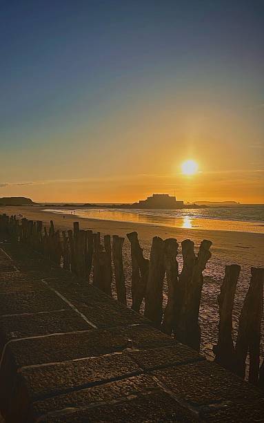 Golden hour at Saint-Malo, with the sun setting behind the historic fort, casting a warm glow over the beach. The wooden breakwaters stand tall, weathered by time and tides, framing the tranquil seascape.