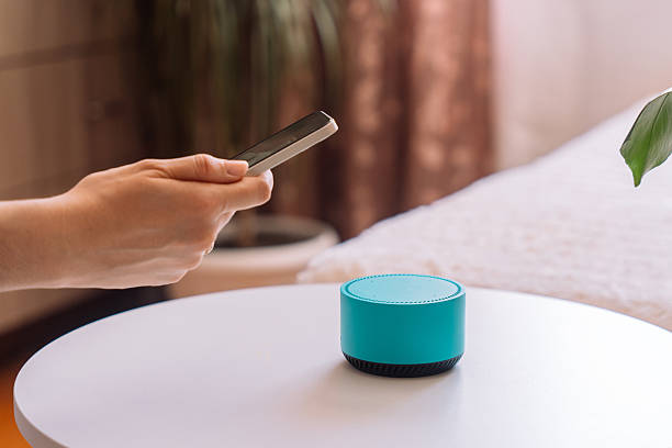 istock Female hand holding a smartphone in front of a blue smart speaker on a white table 2212892452