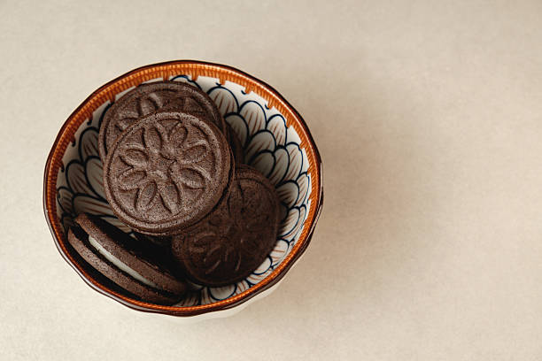 A charming bowl holds an assortment of dark, round cookies, showcasing intricate designs. The bowl's vivid patterns enhance the allure of the delicious snacks against a minimalist background.