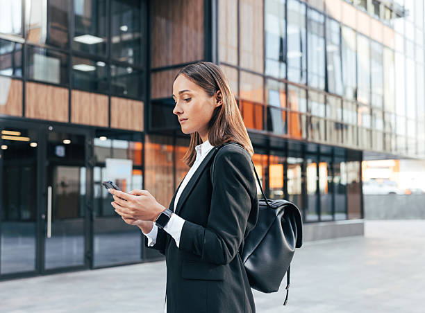 istock Female in black formal clothes wearing backpack typing on mobile phone while standing against an office building 2212906727