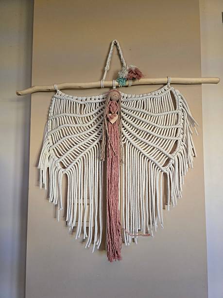 istock A beautiful macrame wall hanging, crafted from white and pink cords, features a driftwood branch at its top, showcasing intricate knots. 2212917518