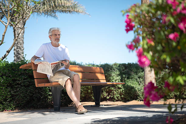 An elderly man sits on a bench in a park, deeply engrossed in reading a newspaper. The vibrant flowers in the foreground and clear sky create a serene atmosphere.