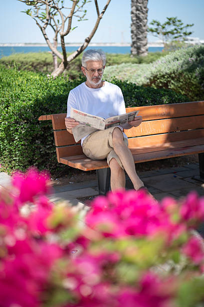 A man, deeply focused on reading a newspaper, sits on a bench near the waterside. The surrounding greenery and flowers add to the peaceful outdoor setting.