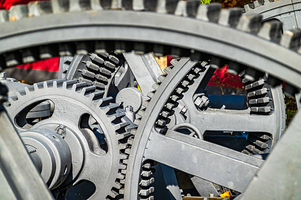 Close-up of large metallic gears engaged in a mechanical process, highlighting intricate design and functionality. Sunlight casts shadows, emphasizing the textures and precision of the machinery.