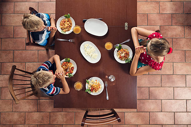 Three kids are gathered around a wooden table, praying before the meal. The overhead view emphasizes the shared meal and togetherness, highlighting the interactions between the family members. Various dishes, including a plate of mixed vegetables and a bowl of rice, are arranged on the table. The tiled terracotta floor beneath the table is typical to the interior in Tuscany, Italy.\n\nShot with Nikon D850