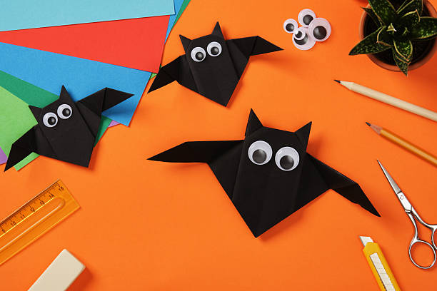 istock Three handmade black origami bats with googly eyes on a bright orange background surrounded by colorful paper sheets and crafting tools 2212937790