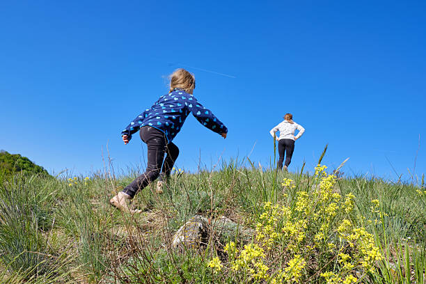 A young child enthusiastically climbs a grassy hillside filled with wildflowers, reaching toward a parent waiting at the top under a clear blue sky. This dynamic image conveys adventure, resilience, and the joy of family exploration in nature.