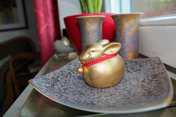 This elegant photo features a round golden bunny figurine with a red collar and small bell, resting on a handcrafted ceramic plate. The setting is complemented by luxurious vases with gold accents and a cozy background of soft red and earthy tones. The composition exudes festive charm and tasteful design, making it ideal for Easter, interior styling, or lifestyle and decoration themes.