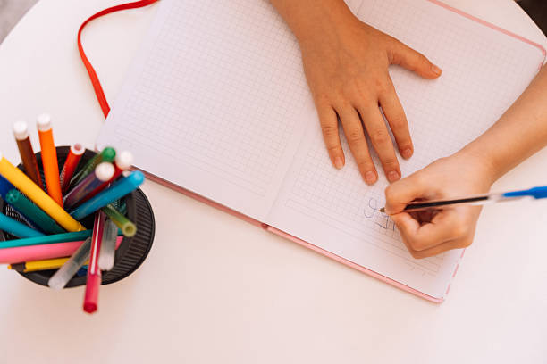 The girl writes with a pen with her left hand in a notepad on a white table.