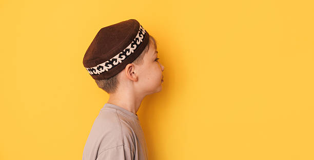 A boy in a Kazakh skullcap stands sideways to a yellow background.