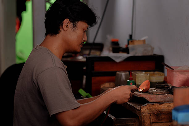 A focused male artisan works over a rustic bench, melting metal in a clay crucible using a handheld torch. Surrounded by tools and containers, the scene captures the dedication and environment of traditional metalworking craftsmanship.
