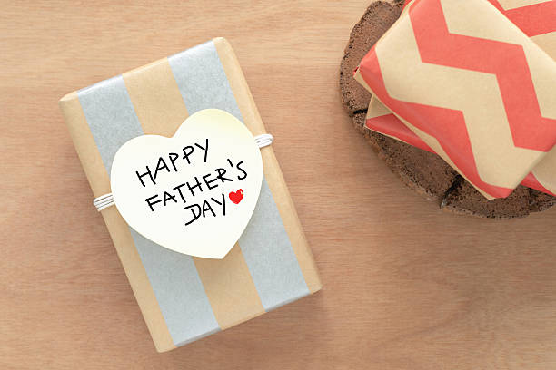 Top view, gift box rectangular wrapped in alternating vertical stripes of  brown and silver paper. A white heart-shaped tag is handwritten with the words HAPPY FATHER'S DAY and a small red heart.