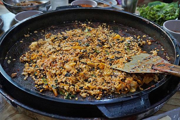 A savory Korean fried rice dish known as bokkeumbap, prepared in a large pan and packed with bold flavors. The rice is stir-fried with various vegetables, possibly including kimchi for an added tangy depth. Fresh greens sprinkled on top enhance the color and texture, making it visually appealing. Surrounding the pan are multiple bowls and plates, suggesting a communal meal, which is a signature aspect of Korean dining culture. The dish is typically enjoyed with a side of soup, pickled vegetables, and dipping sauces.