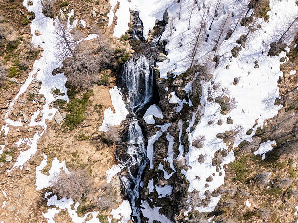 Dramatic aerial shot of a waterfall cascading down a snowy mountain slope. Melting snow and rocky terrain create a stunning contrast. Nature transitioning from winter to spring.