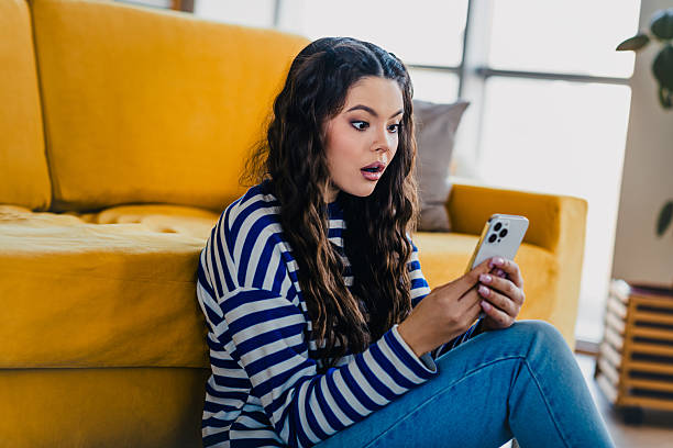 istock Young woman in casual striped pullover surprised by smartphone message while relaxing on couch in bright living room 2213011661