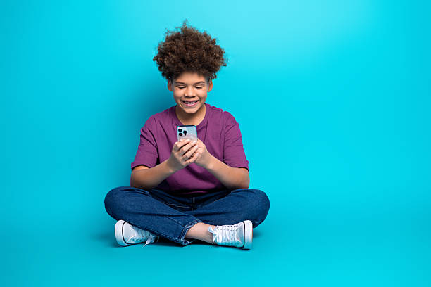 istock Smiling boy with curly hair using smartphone on a bright blue background, showcasing trendy casual fashion and joyful emotions 2213013806