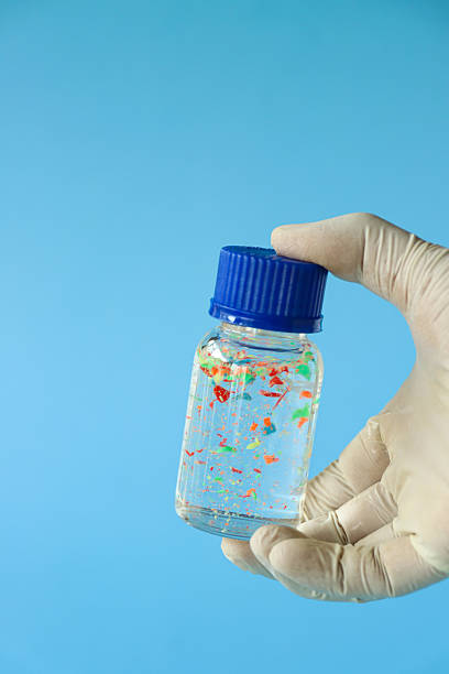 Image of multicoloured flakes and chips of plastic suspended in water, sample bottle held between tub and index finger, unrecognisable person wearing medical gloves, blue background, focus on foreground, microplastics concept, copy space stock photo