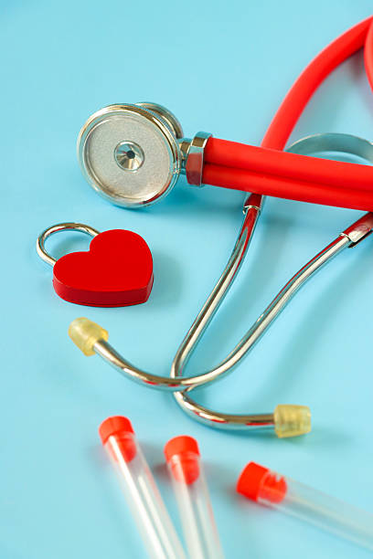 Stock photo showing close-up, elevated view of a stethoscope with vials and a red heart-shape  padlock on a blue background.