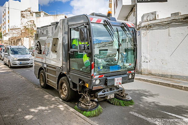 Arrecife, Lanzarote, Canary Islands, Spain - March 10th 2025: The image shows a municipal street sweeper vehicle operating on a city street. The vehicle is equipped with large rotating brushes at the front, designed to clean the road surface efficiently. It is painted in a combination of grey and black, with red and white reflective stripes for visibility. The brand name \