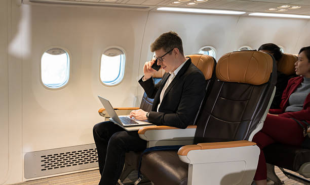 istock Cheerful businessman in black suit using laptop computer while talking on cellphone in airplane cabin 2213017389