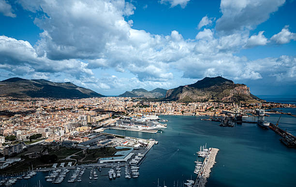 A stunning panoramic view of Palermo's cityscape with its picturesque harbor, surrounded by vibrant mountains and clear blue skies with fluffy clouds, showcasing natural beauty and Mediterranean charm.
