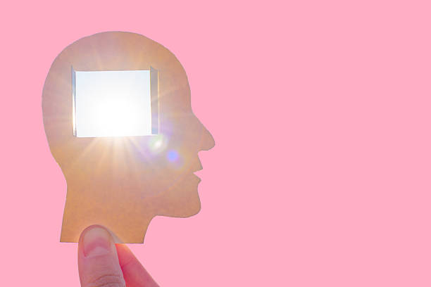Hand holds a paper silhouette of a human head with a window cut-out, allowing bright light to shine through against a pastel pink background with copy space.
