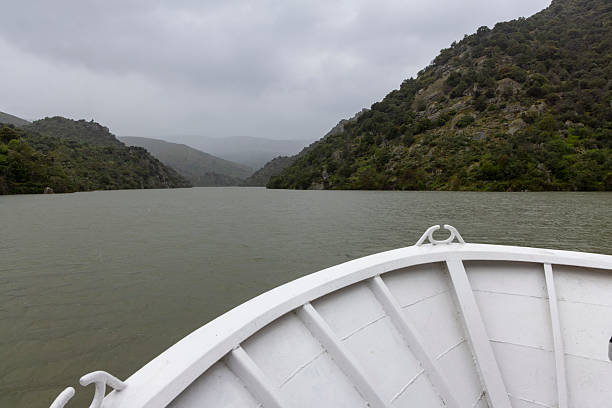 A tranquil boat ride on a river flanked by lush, rugged hills under a cloudy sky. The mist rolls in to create a peaceful ambiance.