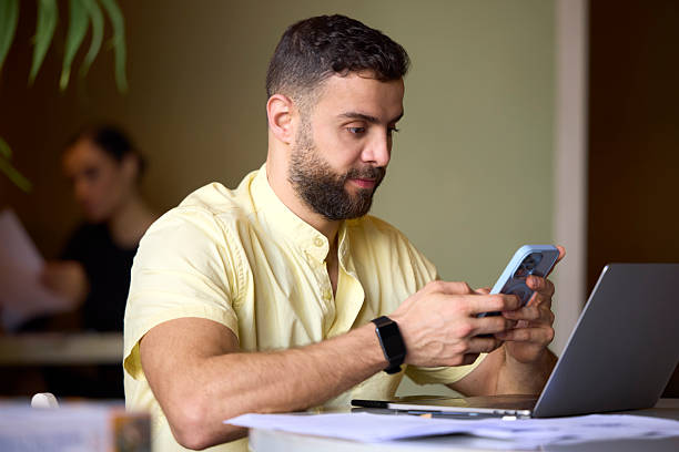Male freelancer using smart phone while sitting at desk in coworking space. Entrepreneur is text messaging at office. He is wearing yellow shirt.