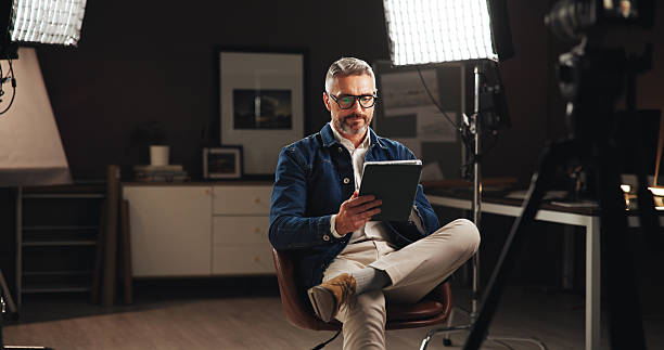 istock Live stream host, recording and man with tablet in home studio for talk show, online interview or podcast. Filming set, lights and mature journalist with digital script for news broadcast or research 2213059079