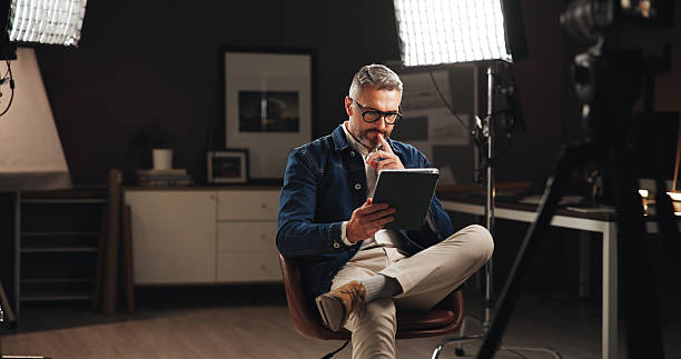 istock Talk show host, man and tablet in home studio for live streaming, online interview and podcast. Filming set, lights and mature journalist with digital script for broadcast research and reading news 2213059086