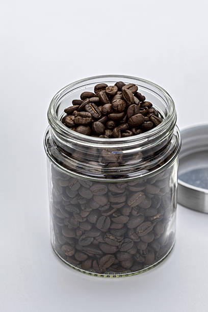 Glass jar filled with aromatic coffee beans, with metal lid placed beside it on white background. image captures rich texture and dark color of beans