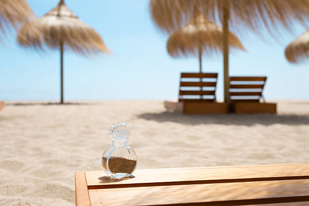 Small glass bottle filled with sand rests on wooden surface on beach with straw umbrellas and deckchairs in the background, creating summer vacation idyllic scene