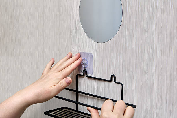 Hands are mounting black wire bathroom shelf on wall using transparent nano adhesive hook for easy and damage-free bathroom hardware fitting